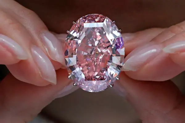 Dłonie trzymają olśniewający, owalny, różowy diament, prawdopodobnie najdroższy diament na świecie, błyszczący w świetle.